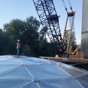 Worker in a mask on top of a large geodesic tank dome with boom lift in background