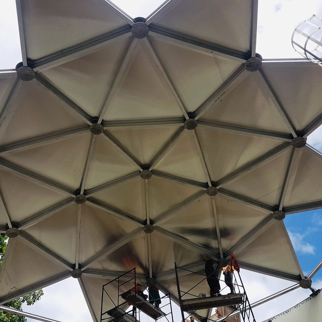 Underside of an aluminum geodesic dome in a star shape. Cloudy blue sky and safety cage.