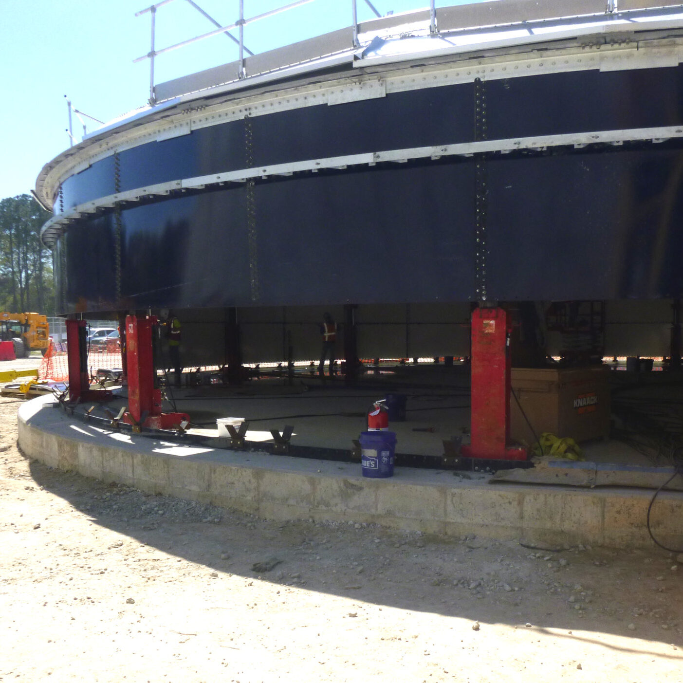Red hydraulic jacks lifting steel water tank panels at a jobsite.