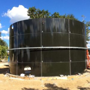Dark green bolted water tank with exposed nozzles. Blue sky, dirt lot.
