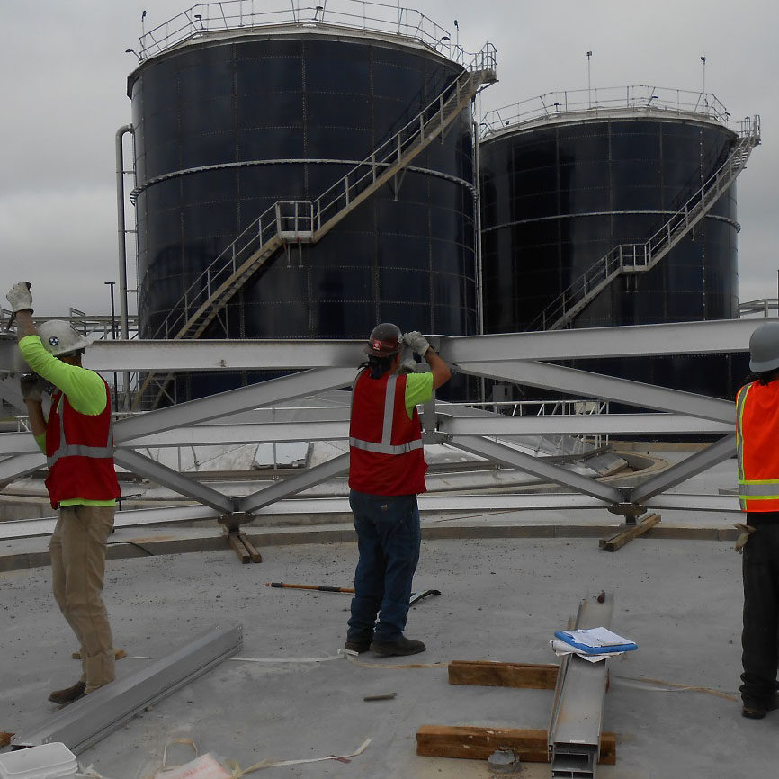 Installers erecting the beams for an aluminum dome tank roof. Two bolted tanks behind.