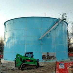 Light blue bolted tank with a stairwell being installed on the side. Small green bobcat in the front.