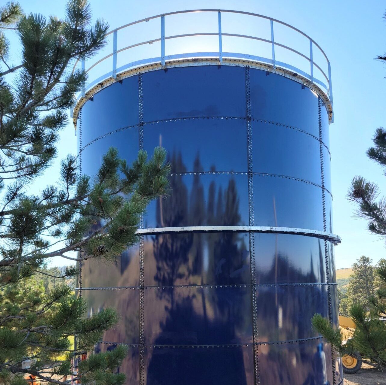 Blue water storage tank with safety rail. Pine trees in front