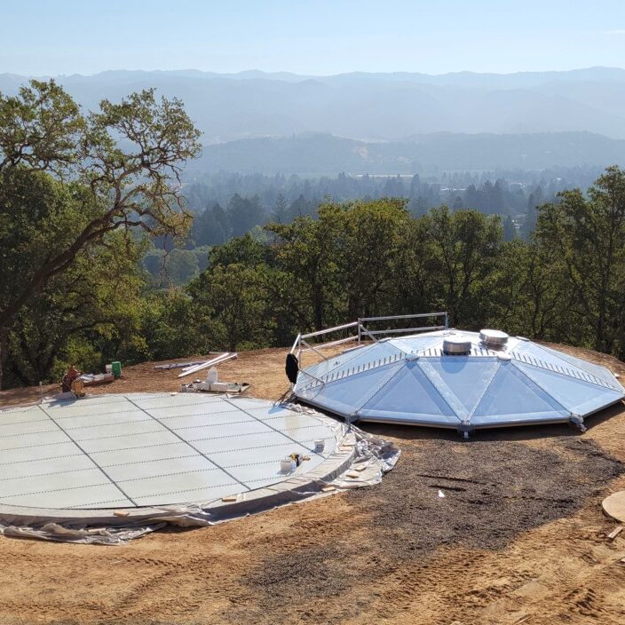 Tank floor and aluminum dome laying atop a dirt hill. Trees and mountains in the background. Blue Sky.