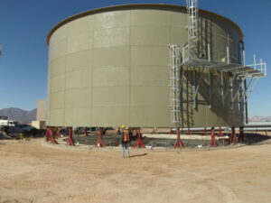Tan colored bolted water storage tank with aluminum platform and red hydraulic jacks. Worker in front