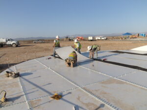 Workers bolted sheets of metal to bolted tank floor. Mountains in background