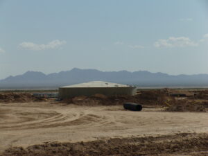 Tank water tank with aluminum dome. Black pipe in foreground with dirt road. Mountains in background
