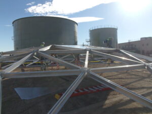 Worker on top of aluminum geodesic dome structure with two water storage tanks and a building in background