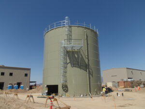 Worker leaving through opening in a bolted storage tank. Caged ladder and platform in foreground. Two buildings in background. Blue Sky and safety rails.