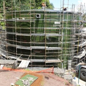 Scaffolding surrounding a circular green metal water tank with installers on top of the tank roof.