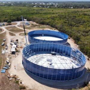 Two three million gallon blue water storage tanks being erected on a dirt pad on a construction site surrounded by green vegetation.